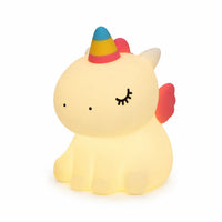 Veilleuse en silicone en forme de petite licorne assise, allumée sur un fond blanc uni. Le personnage présente des yeux clos avec des cils dessinés, une corne colorée en dégradé de bleu, jaune et rose, et de petites ailes roses, diffusant une douce lueur chaude et tamisée.
