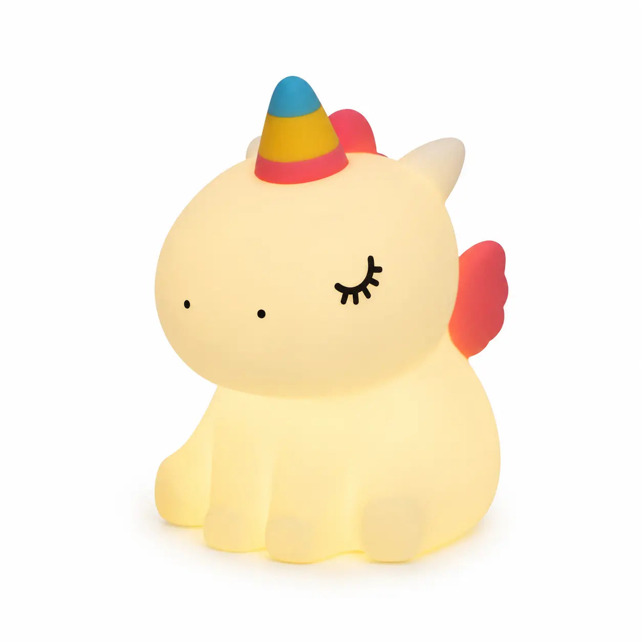 Veilleuse en silicone en forme de petite licorne assise, allumée sur un fond blanc uni. Le personnage présente des yeux clos avec des cils dessinés, une corne colorée en dégradé de bleu, jaune et rose, et de petites ailes roses, diffusant une douce lueur chaude et tamisée.