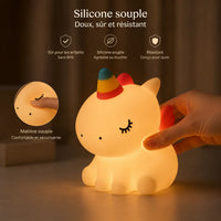 Infographie en français mettant en avant la souplesse d'une veilleuse licorne en silicone allumée. Une main presse doucement le dos du produit pour montrer sa flexibilité, et un insert circulaire à gauche zoome sur un doigt enfonçant sa tête avec la mention "Matière souple confortable et sécurisante". Le haut de l'image présente trois pictogrammes détaillant ses propriétés : sûr pour les enfants sans BPA, agréable au toucher, et résistant.