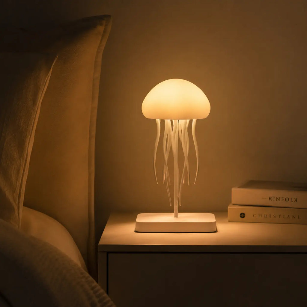 Veilleuse en silicone en forme de méduse allumée sur une table de chevet, diffusant une douce lueur chaude à côté de livres empilés et d'un lit confortable dans une atmosphère de nuit tamisée.