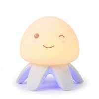Veilleuse en silicone en forme de petite méduse mignonne allumée, avec un dôme souple diffusant une lueur jaune chaleureuse, un visage souriant qui fait un clin d'œil, et des tentacules blancs rétroéclairés par une lumière bleue à la base, présentée sur fond blanc.