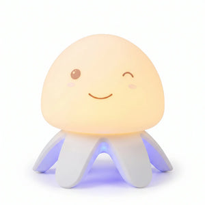 Veilleuse en silicone en forme de petite méduse mignonne allumée, avec un dôme souple diffusant une lueur jaune chaleureuse, un visage souriant qui fait un clin d'œil, et des tentacules blancs rétroéclairés par une lumière bleue à la base, présentée sur fond blanc.