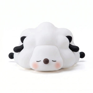 Veilleuse en silicone en forme de petit mouton blanc et noir couché aux yeux fermés, sur un fond blanc.