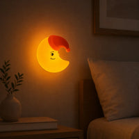 Veilleuse murale en silicone en forme de croissant de lune jaune allumée, fixée sur le mur d'une chambre au-dessus d'un lit. Elle diffuse une douce lueur orange tamisée à côté d'un oreiller, d'un cadre et d'une petite plante en pot sur une table de chevet, créant une ambiance calme et propice au sommeil.