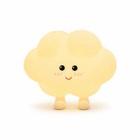 Veilleuse en silicone en forme de petit nuage jaune pâle tout rond doté de deux petits pieds. Elle présente un visage joyeux de style kawaii dessiné de face avec des yeux ronds noirs, de petites pommettes rosées et un grand sourire rouge, présentée sur un fond blanc épuré.