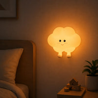 Veilleuse murale en silicone en forme de petit nuage à pieds allumée, fixée au mur. Elle diffuse une douce lueur chaude orangée au-dessus d'une table de chevet dans une chambre d'enfant à l'ambiance cocooning.