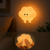 Veilleuse murale en silicone en forme de petit nuage à pieds allumée au mur, diffusant une douce lueur chaude orangée à côté d'un lit. Un zoom circulaire au bas de l'image montre le produit en silicone souple pressé doucement entre les doigts.