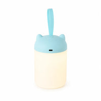 Veilleuse nomade rechargeable dotée d'une anse de transport suspendue et d'un couvercle bleu ciel à petites oreilles, montrant le port USB de recharge et la base cylindrique illuminée d'une lueur chaude sur fond blanc.