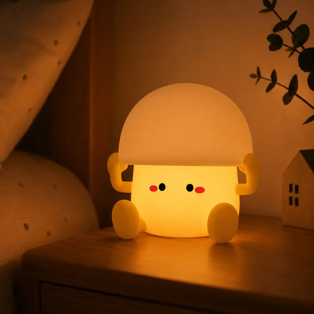 Veilleuse œuf à la coque en silicone allumée diffusant une lueur chaleureuse et tamisée sur une table de chevet en bois. Elle est installée dans une chambre à coucher à côté d'un oreiller beige et d'une petite décoration en forme de maison.