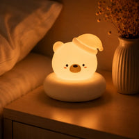 Veilleuse blanche en forme de tête de petit ours allumée, posée sur son large socle circulaire de couleur crème sur une table de chevet en bois à côté d'un lit. Portant son bonnet de nuit incliné sur le côté, le personnage diffuse une lueur chaude et tamisée dans une chambre à coucher à l'ambiance douce et apaisante, juste à côté d'un vase blanc contenant des fleurs séchées.