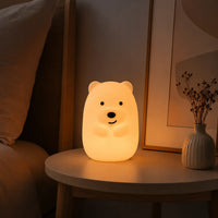 Veilleuse en forme de petit ours allumée posée sur une table de chevet à côté d'un vase dans une ambiance chaleureuse.