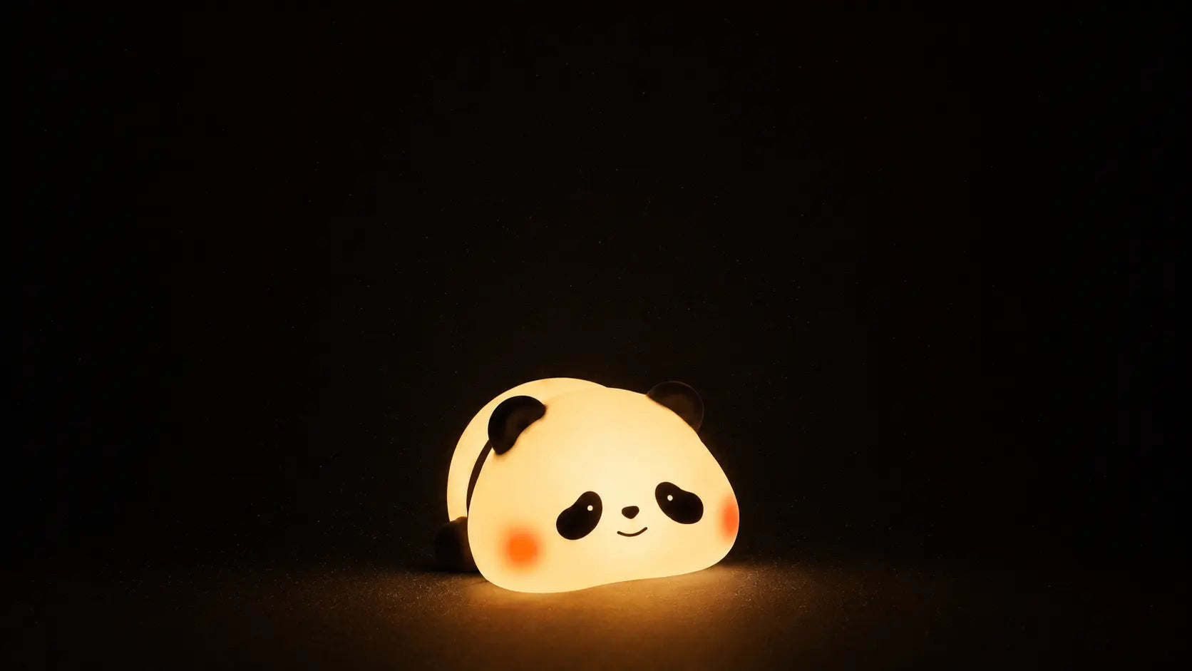 Veilleuse nomade en silicone en forme de petit panda souriant, allumée et diffusant une lumière chaude tamisée au milieu d'une pièce plongée dans le noir complet.