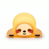 Veilleuse en silicone en forme de petit paresseux allongé tout mignon. Elle affiche un visage serein avec des yeux clos souriants, des taches marron caractéristiques et de petites griffes noires, diffusant une douce lueur jaune tamisée sur un fond blanc épuré.