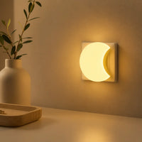 Veilleuse en forme de lune branchée sur une prise murale, diffusant une lumière chaude et apaisante à côté d'un vase décoratif.