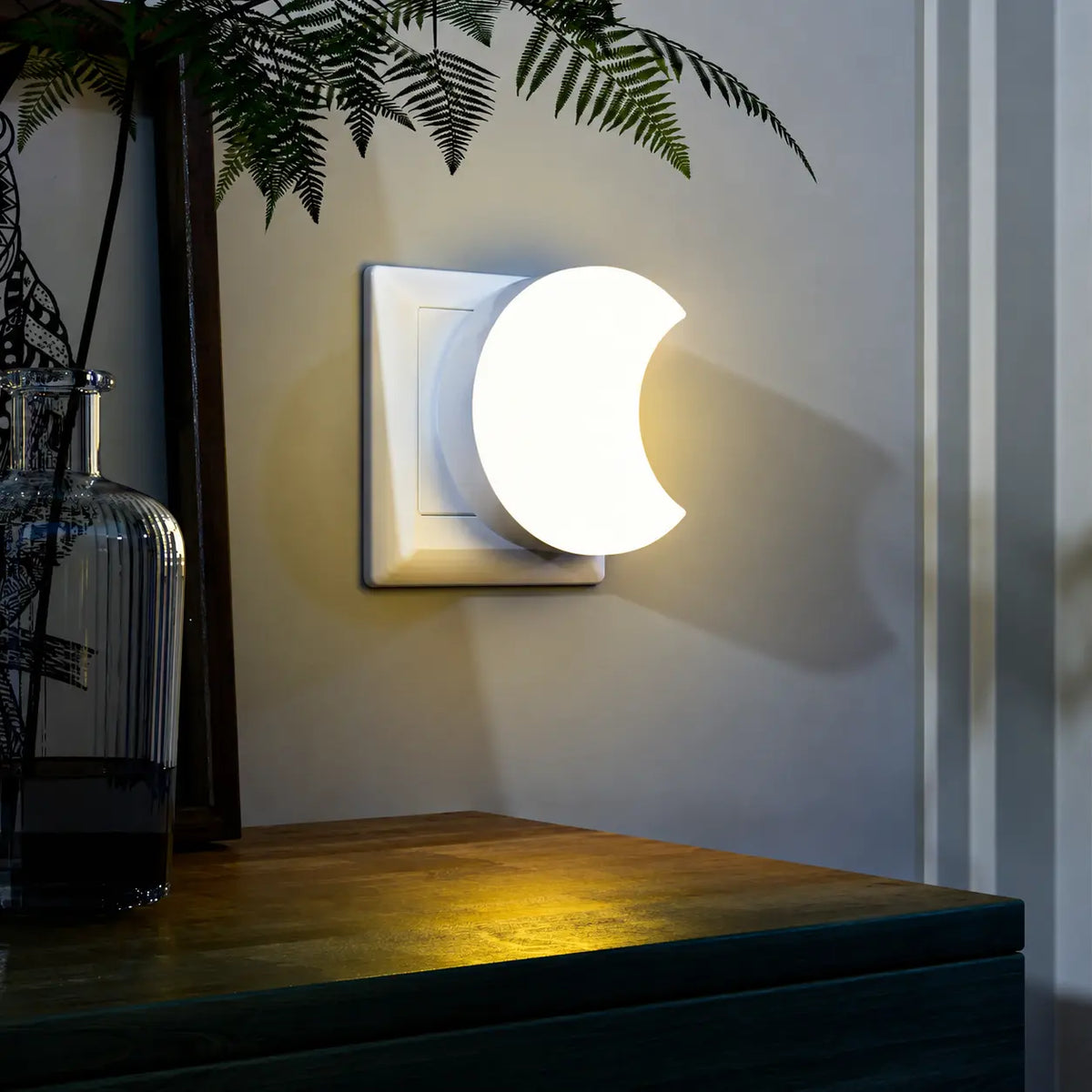 Veilleuse lune murale illuminée au-dessus d'un meuble en bois sombre avec des plantes d'intérieur dans une atmosphère de soirée.