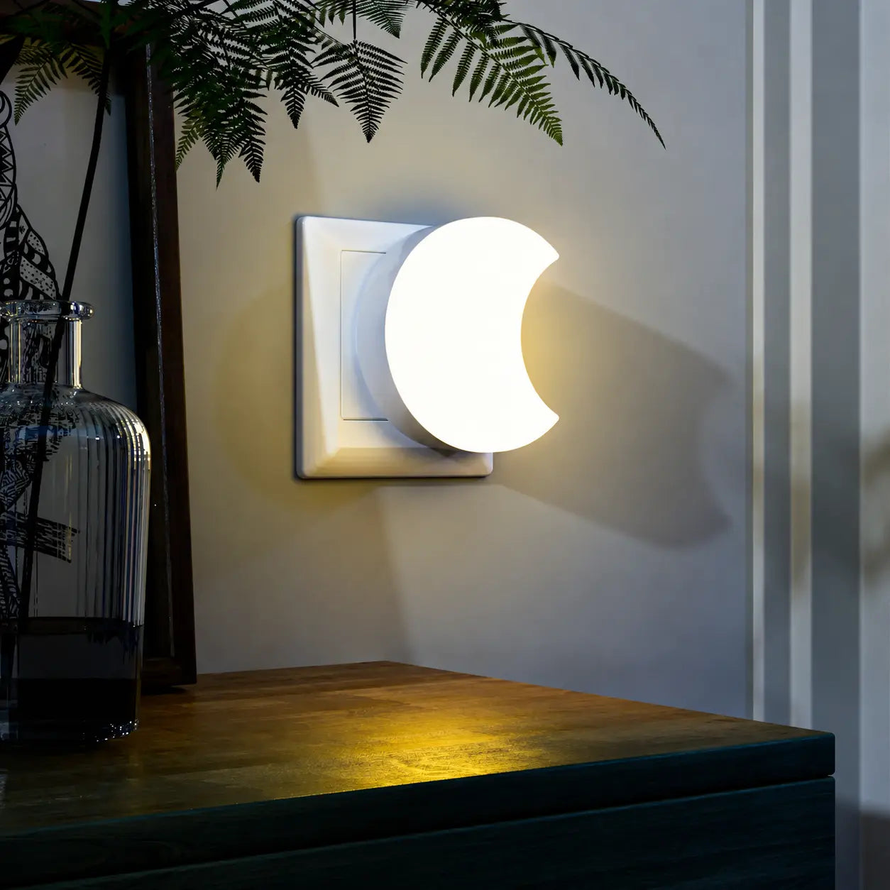 Veilleuse lune murale illuminée au-dessus d'un meuble en bois sombre avec des plantes d'intérieur dans une atmosphère de soirée.