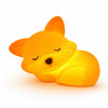 Veilleuse en silicone en forme de petit renard endormi allumée, recroquevillée et diffusant une lueur orange et jaune chaleureuse, avec des yeux clos dessinés, un petit museau noir et des détails gravés sur le flanc, présentée de face sur fond blanc.