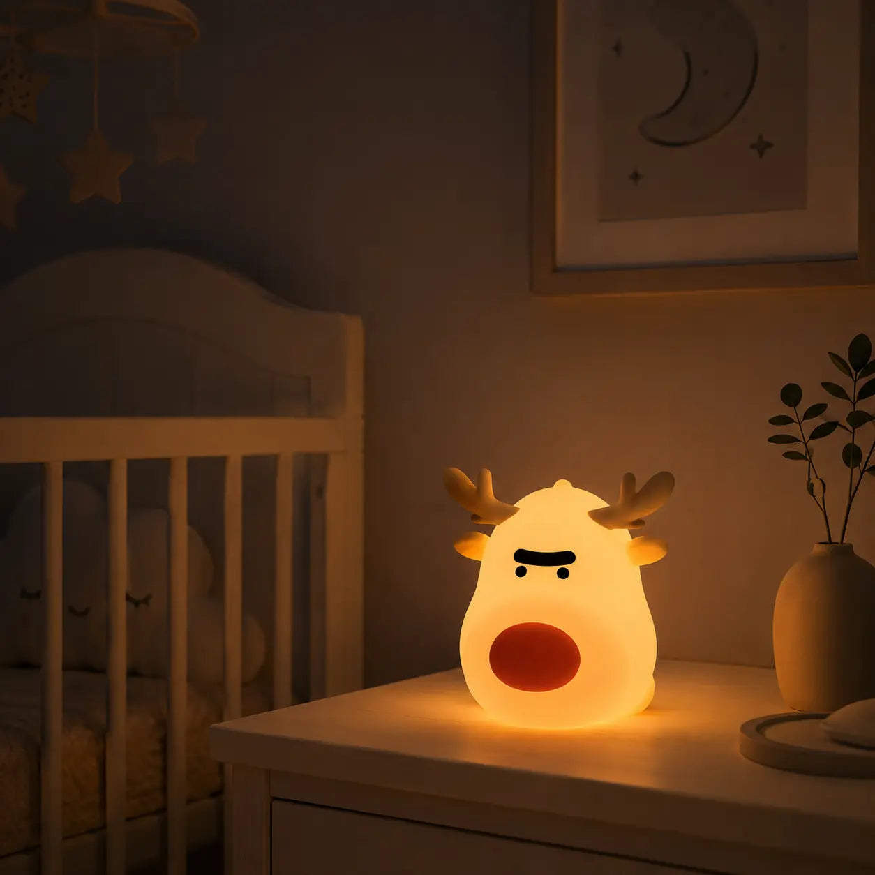 Veilleuse renne au nez rouge allumée sur une commode en bois blanc dans une chambre d'enfant. Elle diffuse une douce lueur tamisée à côté d'un vase en céramique et d'un lit à barreaux blanc en arrière-plan, sous un tableau illustrant la lune.