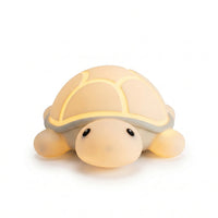 Veilleuse nomade en silicone souple en forme de petite tortue mignonne, allumée et diffusant une douce lueur chaude et tamisée à travers sa carapace, présentée de face sur fond blanc.