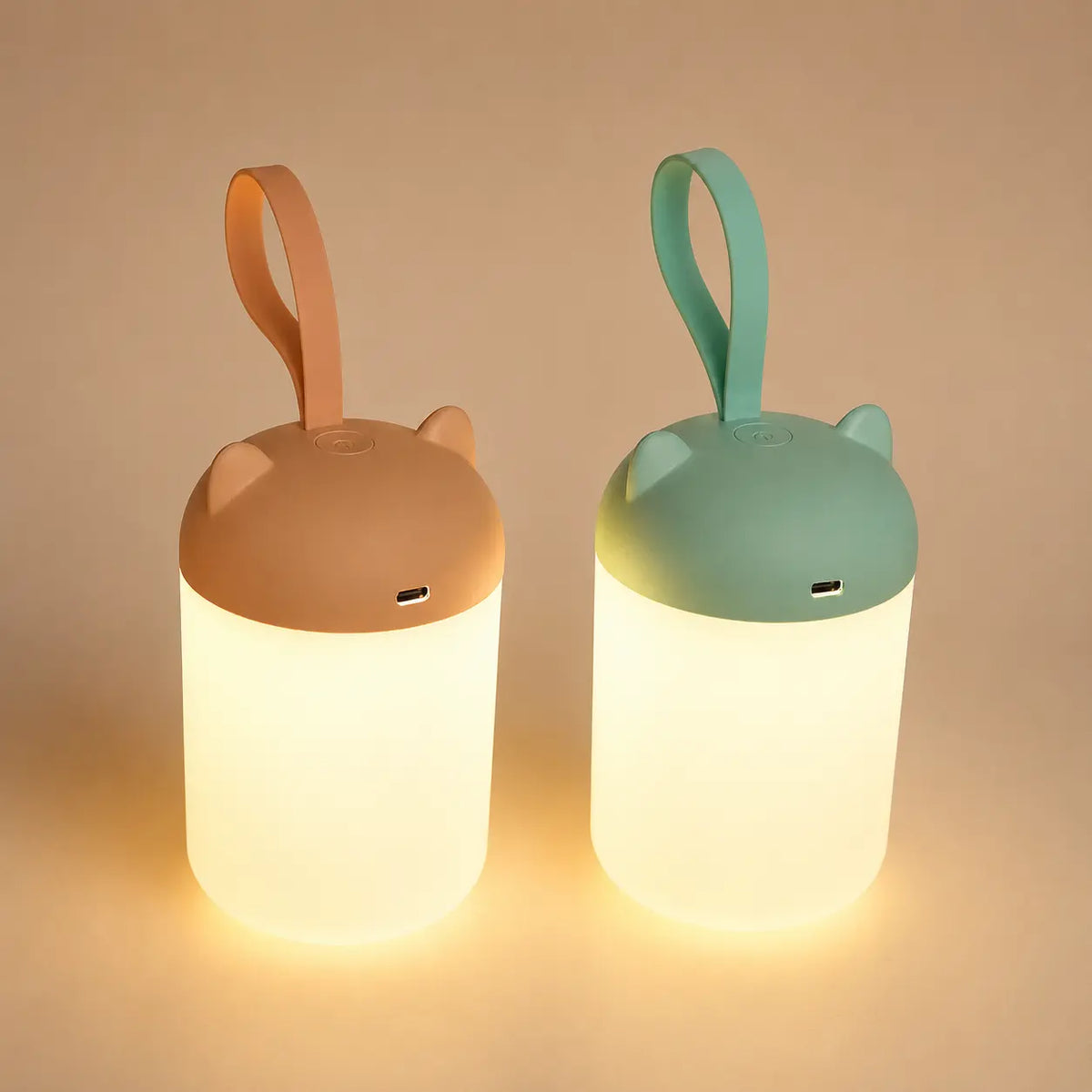 Deux veilleuses nomades à anse allumées côte à côte, l'une couleur terracotta et l'autre vert sauge, diffusant une douce lueur chaude sur un fond épuré.