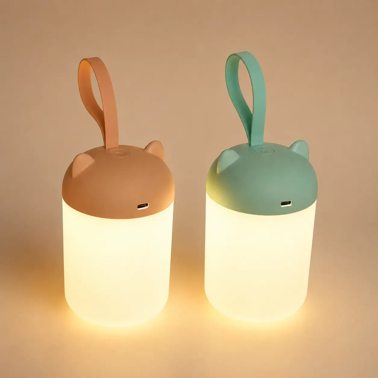 Deux veilleuses nomades à anse allumées côte à côte, l'une couleur terracotta et l'autre vert sauge, diffusant une douce lueur chaude sur un fond épuré.